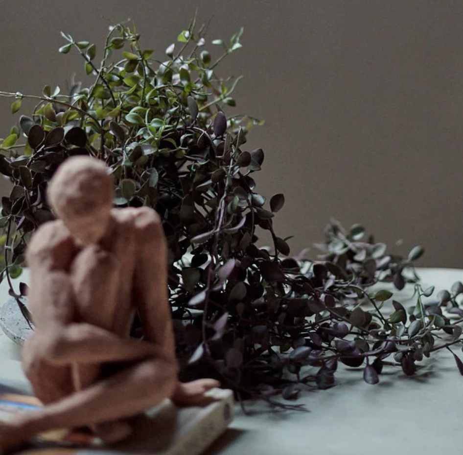 Wooden sculpture of a person sitting among green plants on a neutral background