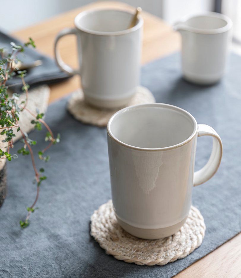 White ceramic mugs on woven coasters on a textured surface with a plant in the background