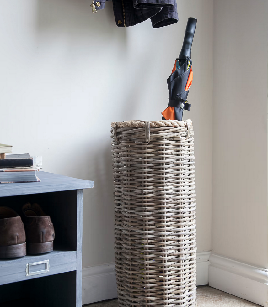 Wicker umbrella stand with an umbrella and shoes on a shelf against a plain wall.