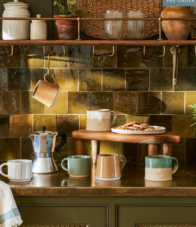 Kitchen counter with ceramic mugs, a coffee maker, and a platter of pastries against a tiled wall.