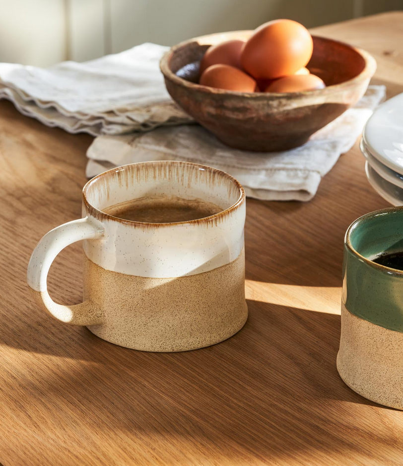 Two ceramic mugs on a wooden table with a bowl of eggs in the background.