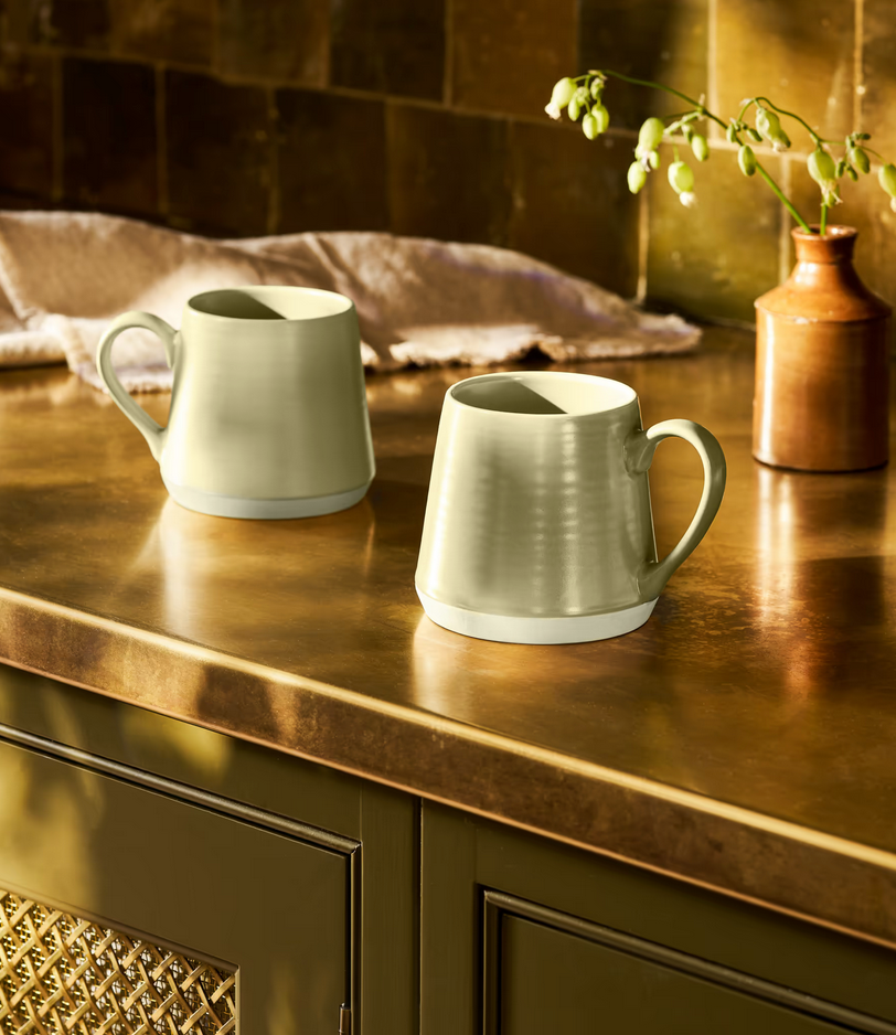 Two ceramic mugs on a wooden surface with a vase in the background.